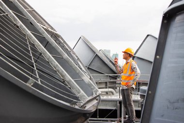 Asyalı erkek mühendis boru tesisatlarını ve soğutucuları inceliyor. Fabrikadaki soğutma tesisi. Kask takan erkek tamirci ve yansıtıcı güvenlik yeleği kondisyon sistemlerinde dizüstü bilgisayar, tablet ve soğutma kuleleri olan ekipman kullanıyor.