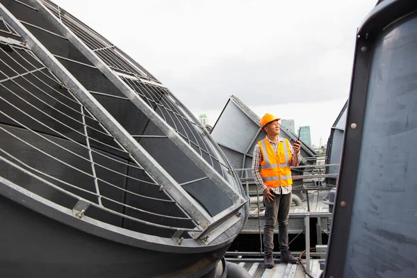 Asian male engineer inspecting pipe fittings and chillers, refrigeration plant refrigeration plant in  factory. male mechanic wearing helmet and Reflective Safety Vest uses laptop, tablet, equipment with sets of cooling towers in conditioning systems