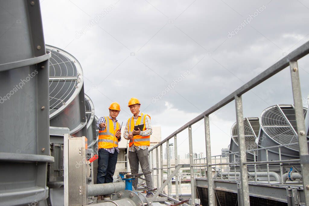 Asian male engineer inspecting pipe fittings and chillers, refrigeration plant refrigeration plant in  factory. male mechanic wearing helmet and Reflective Safety Vest uses laptop, tablet, equipment with sets of cooling towers in conditioning systems