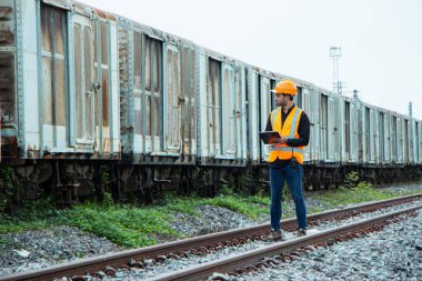Başlık ve yelek giyen Müfettiş Mühendislik demiryolu inşaatını kontrol etmekten aranıyor. Asyalı işçiler ve iş arkadaşları eski tren araçlarının yanında dururlar..