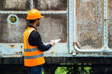 Başlık ve yelek giyen Müfettiş Mühendislik demiryolu inşaatını kontrol etmekten aranıyor. Asyalı işçiler ve iş arkadaşları eski tren araçlarının yanında dururlar..