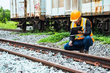 Başlık ve yelek giyen Müfettiş Mühendislik demiryolu inşaatını kontrol etmekten aranıyor. Asyalı işçiler ve iş arkadaşları eski tren araçlarının yanında dururlar..