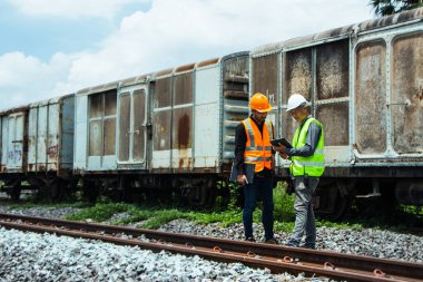 Başlık ve yelek giyen Müfettiş Mühendislik demiryolu inşaatını kontrol etmekten aranıyor. Asyalı işçiler ve iş arkadaşları eski tren araçlarının yanında dururlar..