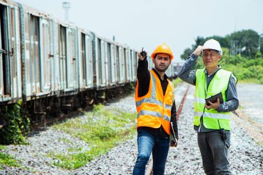 Başlık ve yelek giyen Müfettiş Mühendislik demiryolu inşaatını kontrol etmekten aranıyor. Asyalı işçiler ve iş arkadaşları eski tren araçlarının yanında dururlar..
