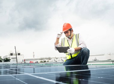 Erkek mühendis bir fotovoltaik (solar) bitkiyi kontrol eder ve bir kayıt tableti kullanır. Koruyucu kasklı tamirci. Üniformalı bir adam tablet tutuyor..