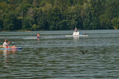 Brno, Çek Cumhuriyeti - 16 Ağustos 2025 - Brno Barajı - İnsanlar güneşli bir günde yemyeşil ağaçlarla çevrili sakin bir gölde çeşitli su sporlarının tadını çıkarıyorlar