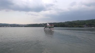 Brno, Çek Cumhuriyeti - 16 Ağustos 2025 - Brno Barajı - uzak yelkenlileri ve ağaçlarla kaplı bir kıyısı olan bir barajda tur teknesi