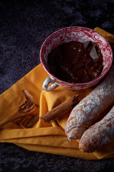Fartons, typical pastries from Valencia, Spain, with hot chocolate, in a Chinese ceramic container on top of a yellow cloth, with a dark surface. A sweet breakfast or snack.