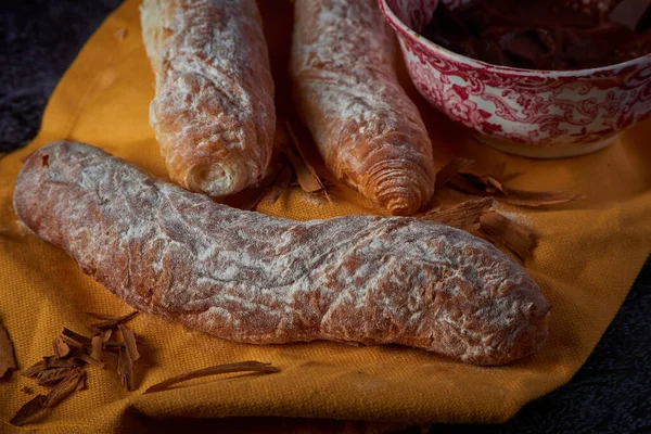 Fartons, typical pastries from Valencia, Spain, with hot chocolate, in a Chinese ceramic container on top of a yellow cloth, with a dark surface. A sweet breakfast or snack.