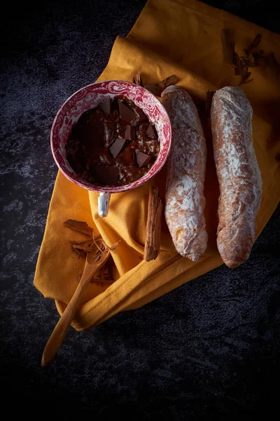 Fartons, typical pastries from Valencia, Spain, with hot chocolate, in a Chinese ceramic container on top of a yellow cloth, with a dark surface. A sweet breakfast or snack.