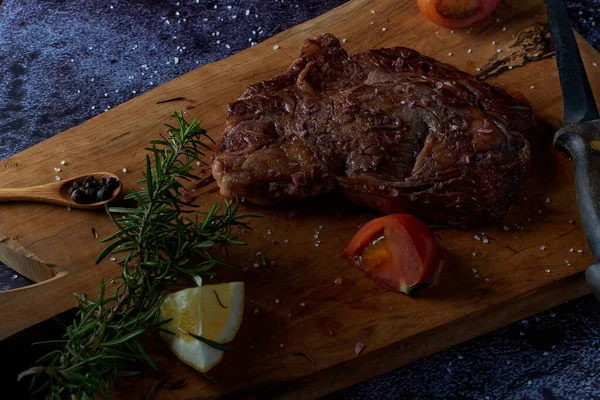 Grilled beef steak on wooden board with salt and other seasonings on a dark surface. Steak ready for eating. Dark food. Meat, tomato, pepper, lemon and rosemary.