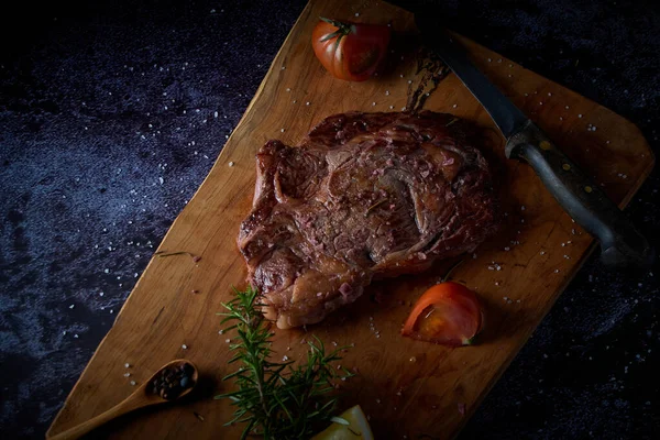 Grilled beef steak on wooden board with salt and other seasonings on a dark surface. Steak ready for eating. Dark food. Meat, tomato, pepper, lemon and rosemary.