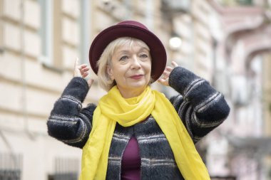 60-65 yaşlarında yaşlı bir kadın, şehir manzarasının arka planında şık bir şapka ayarlıyor. Kavram: Emeklilik, turizm ve seyahat, ileri yaş modası, mükemmel sağlık.