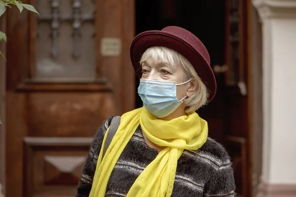 An elderly woman 60-65 years old in a medical mask comes out of the entrance of an old house. Concept, retirement, fashionista of advanced years, excellent health, meeting on the street, quarantine and virus, isolation and communication,