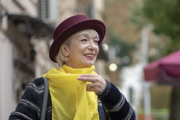 An elderly woman 60-65 years old in a hat adjusts a yellow scarf around her neck against the backdrop of the city landscape. Concept: retirement, fashionista of advanced years, excellent health.
