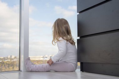 Küçük bir kız kocaman bir pencerenin önünde bir apartmanın zemininde oturuyor ve şehre bakıyor. Kavram: Çocukların yalnızlığı, hayalleri ve gözlemleri.