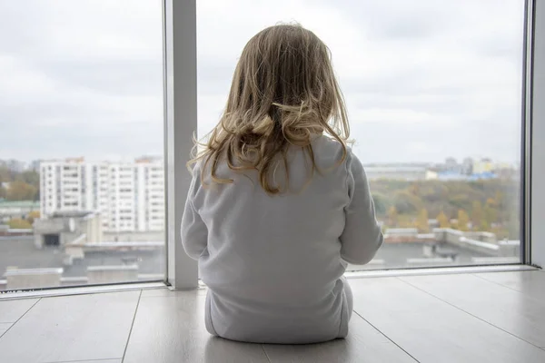 一个小女孩正坐在一个巨大窗户前的公寓里的地板上 儿童的孤独 梦想和观察 — 图库照片
