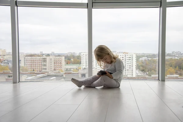 A little girl sits on the floor in front of huge Windows and holds a smartphone in her hands.
