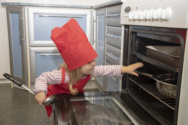 Portrait of a little girl in a chef's hat and apron in the kitchen with a ladle in her hands, opens and looks into the oven.