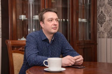 30-35 yaşlarında gülümseyen bir adamın portresi. Masada elinde bir kupayla oturuyor. Evin arka planında. Belki de aktör, TV sunucusu, dişçi ya da avukattır..