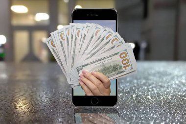 Bir kadının eli cep telefonundan gelen yüz dolarlık banknotları, bulanık bir şehir arkaplanını, yağmur damlalarını tutuyor. Konsept: mobil uygulama, banka kredisi veya piyango kazananı üzerinden para transfer etmek.