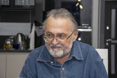 65-70 yaşlarında, gözlüklü yaşlı bir adamın portresi. Mutfakta oturuyor ve kafasını çeviriyor..