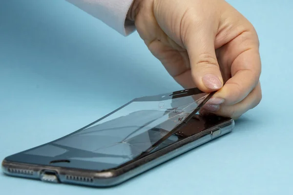 Kadın elleri, mavi arka planda, yakın plan, seçici odak noktasındaki akıllı telefonun kırık koruyucu camını soyar. Kavram: cam değiştirme, telefon tamiratı kendi ellerinle..