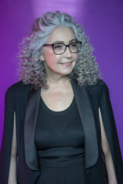 Студийный портрет улыбающейся пожилой женщины 60-65 лет в строгом черном костюме и очках, с седыми кудрявыми длинными волосами, на цветном фоне. Концепция: стильные пенсионеры модельного вида, активная жизнь,