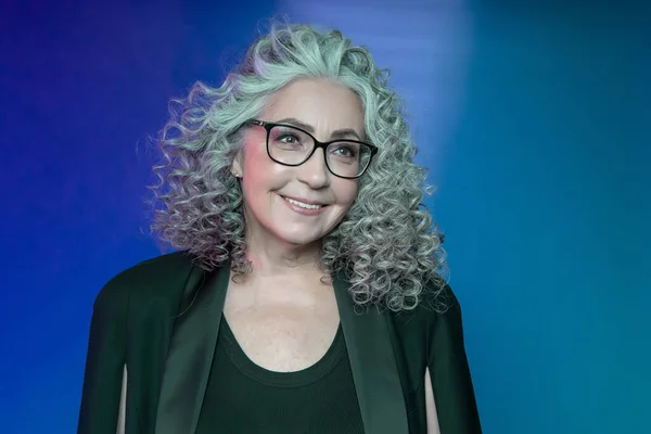 Studio portrait of a smiling elderly woman of 60-65 years old in a strict black suit and glasses, with gray curly long hair, on a colored background. Concept: stylish pensioners of model appearance, active life,