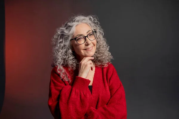 Studio portrait of an elderly woman 60-65 years old in a red sweater and glasses, gray curly long hair, on a colored background. Concept: stylish pensioners of model appearance, active life.