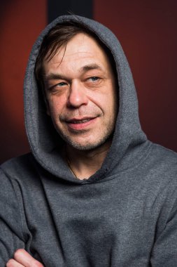 Studio portrait of a 40-50-year-old laughing man in a gray hoodie on a neutral background with a hood on his head, looking away. Maybe he's an actor, or a retired soldier, or a lawyer, or a longshoreman.