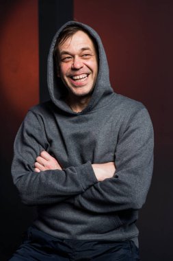 Studio portrait of a 40-50-year-old laughing man in a gray hoodie on a neutral background with a hood on his head, looking away. Maybe he's an actor, or a retired soldier, or a lawyer, or a longshoreman.