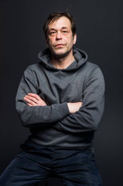 Studio portrait of a 40-50-year-old serious man in a gray hoodie on a neutral background, looking directly at the camera. Maybe he's an actor or a retired military man,