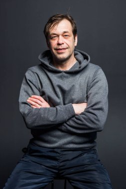 Studio portrait of a 40-50-year-old smiling man in a gray hoodie on a neutral background, looking directly at the camera. Maybe he's an actor or a retired military man,