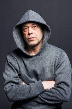 Studio portrait of a 40-50-year-old serious man in a gray hoodie on a neutral background with a hood on his head, looking at the camera, close-up. Maybe he's an actor or a retired military man.