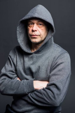 Studio portrait of a 40-50-year-old serious man in a gray hoodie on a neutral background with a hood on his head, looking at the camera, close-up. Maybe he's an actor or a retired military man.