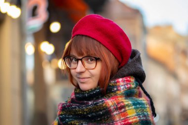 20-25 yaşlarında, gözlüklü, kızıl saçlı ve şehrin arka planına karşı biraz kilolu bir genç kadın. Bir sokak portresinin yakın çekimi. Bereli şişman bir kızın ciddi yüzü..