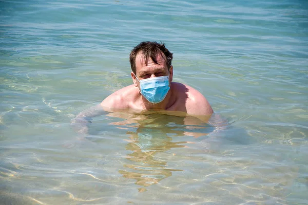 A man of 45-50 years old swims in a medical mask in the sea. Concept: protection from coronavirus, tourist holidays at sea, safety on the beach, opening of the swimming season.