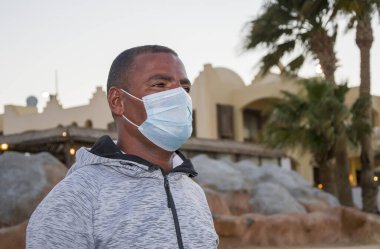 Şehir manzarasının arka planında 40 yıllık tıbbi maske takmış esmer bir adamın portresi. Belki de turist rehberi ya da sahilde cankurtaran olarak çalışıyordur. Menajer ya da hediyelik eşya satıcısı..