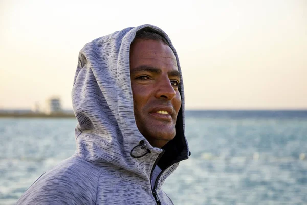 Portrait of a dark-skinned Egyptian man of 40 years in a hoodie against the background of the sea and a boat.