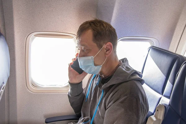 A 45-50-year-old man is sitting in the cabin of an airplane wearing a medical mask and talking on a mobile phone. Concept:violation of the rules of air travel, communication with loved ones during the trip.