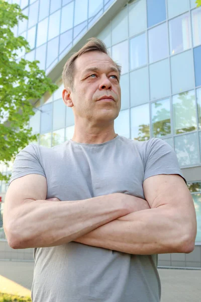 Street portrait of a strong physique man of 40-50 years old in a gray T-shirt against the background of a business center. Concept: job search after retirement, the ability to take out a bank loan.