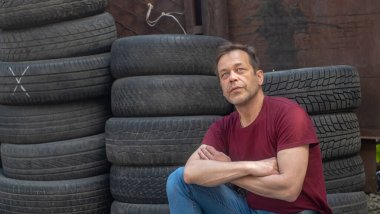 45-50 yaşlarındaki ciddi yaşlı bir adamın portresi eski araba lastiklerinin ve tuğla duvarların arkasında oturuyor. Konsept: Çok çalışma, oto tamirhanesinde çalışma.