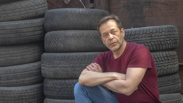 Portrait of a man with a strong build of 45-50 years old against the background of old car tires and a brick wall.Concept: hard work of an immigrant, working in an auto repair shop.
