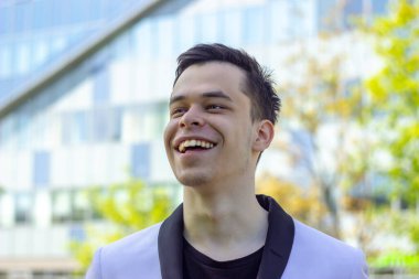 18-20 yaşlarında, ofis binasının arka planında beyaz ceketli, gülen bir gencin sokak portresi. Yakın plan, seçici odaklanma. Konsept: neşe ve dikkatsizlik, eğitimin başlangıcı ya da sonu, iş bulma..
