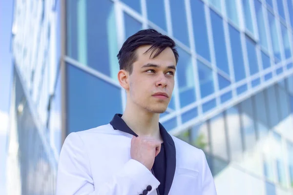 Portrait of a young guy 17-21 years old in a white jacket on the background of glass windows of an office building. Concept: start of training and practice, get a job after college or institute, a new workplace in the office.
