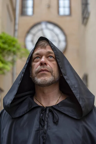 Portrait of an elderly monk 45-50 years old with a beard in a black cassock against the background of old buildings and clocks. Concept: healing and pilgrimage, a journey to holy places.