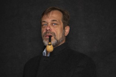 45-50 yaşlarında, siyah ceketli, gri sakallı, siyah arka planda pipo içen ve kameraya bakan yaşlı bir adamın portresi. Kavram: Emekli asker, yazar ya da denizci.