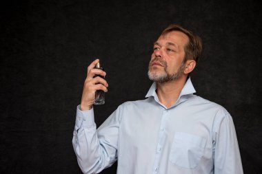 45-50 yaşlarında, beyaz sakallı, açık renk tişörtlü yaşlı bir adam arka plandaki cam şişeden parfüm sıkar..