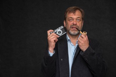 Portrait of an elderly man 45-50 years old with a pipe in his mouth and a black coat on a dark background holding an old film camera.
